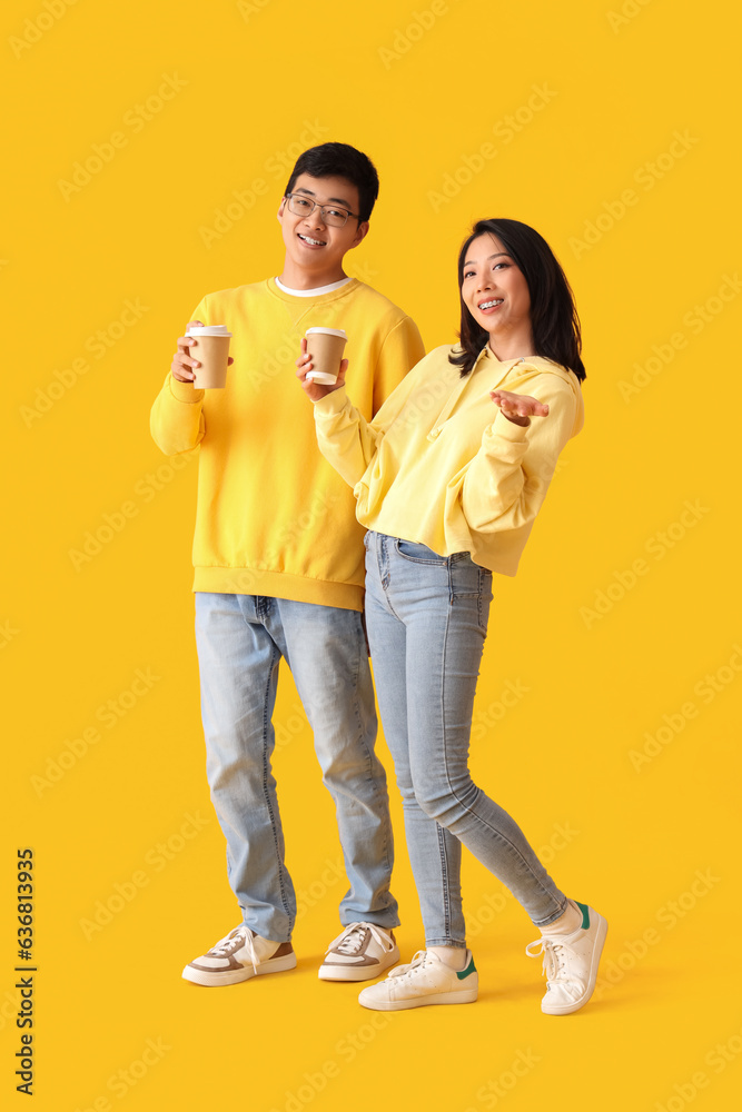 Young Asian friends with cups of coffee on yellow background