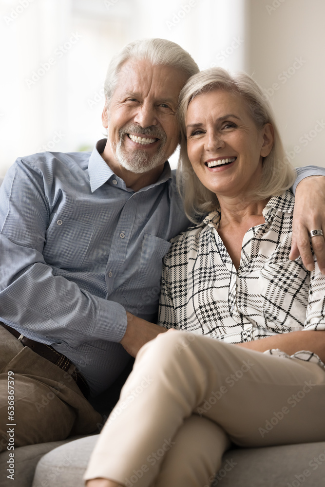 Cheerful positive senior retired husband and wife hugging on home sofa, looking at camera with tooth