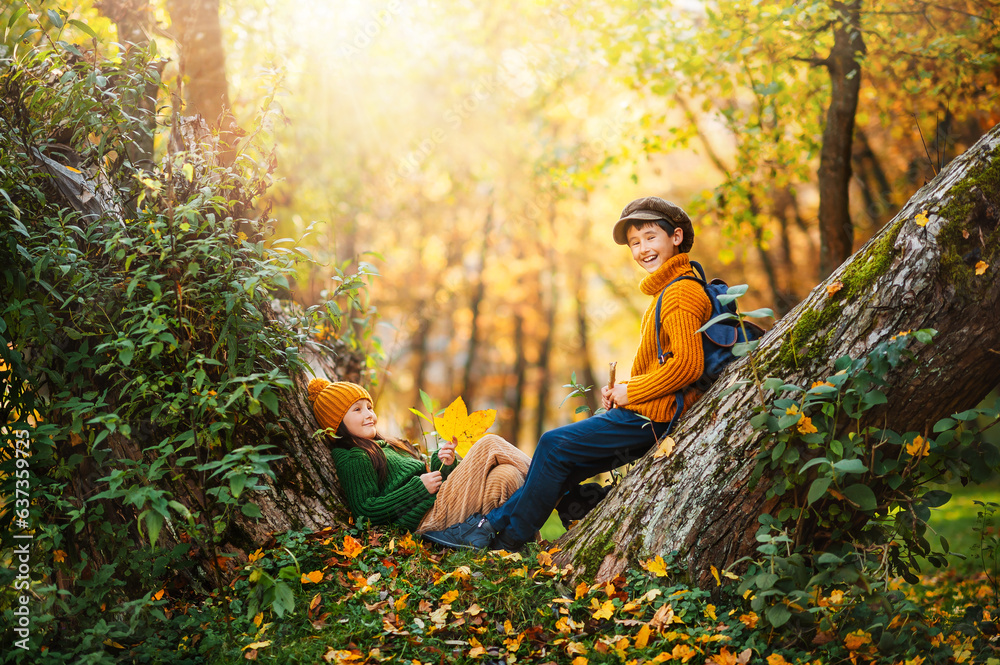 Two happy little children are resting in the autumn park sitting on a dry yellow leaf. Wonderful aut