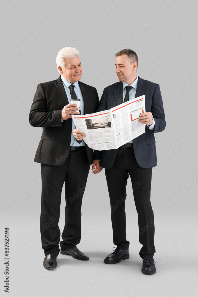 Mature brothers with cup of coffee reading newspaper on grey background