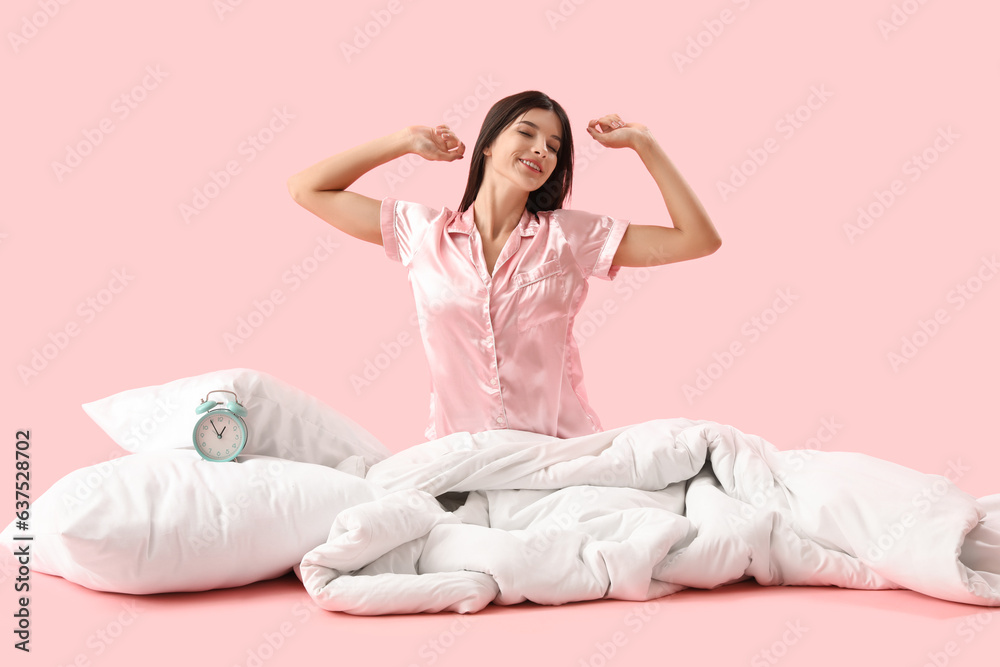 Awaking beautiful young woman with soft blanket and alarm clock on pink background