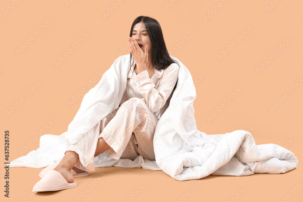 Beautiful young woman with soft blanket yawning on pale orange background