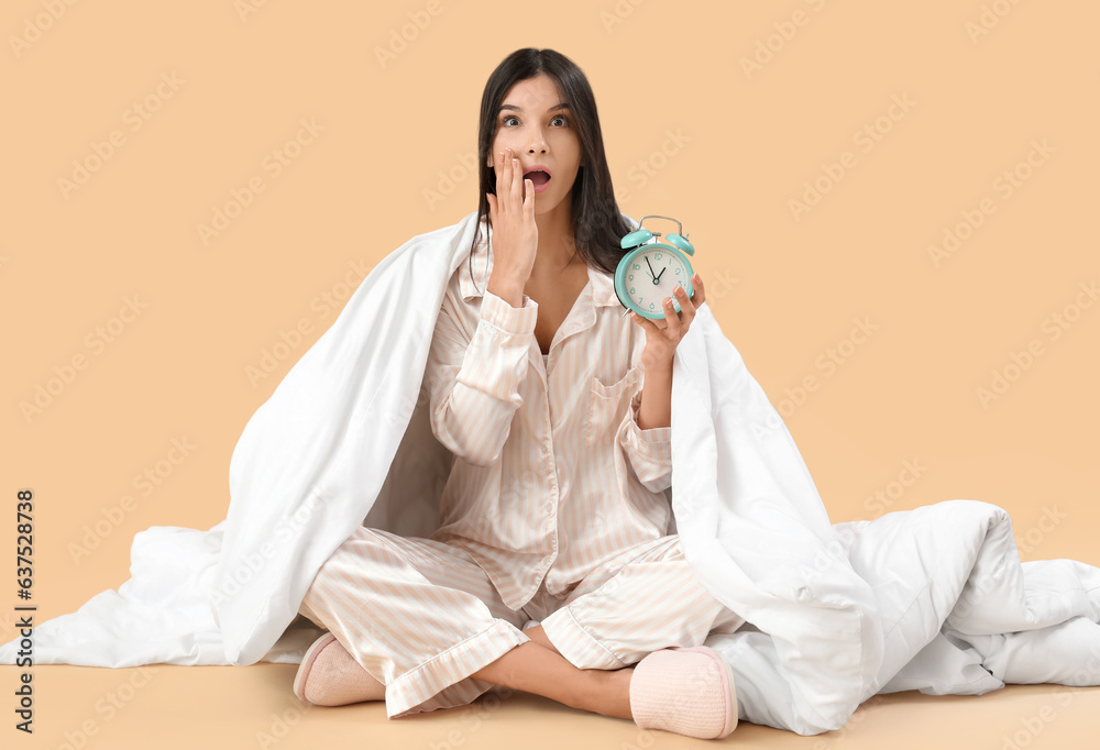 Beautiful shocked young woman with soft blanket and alarm clock sitting on pale orange background