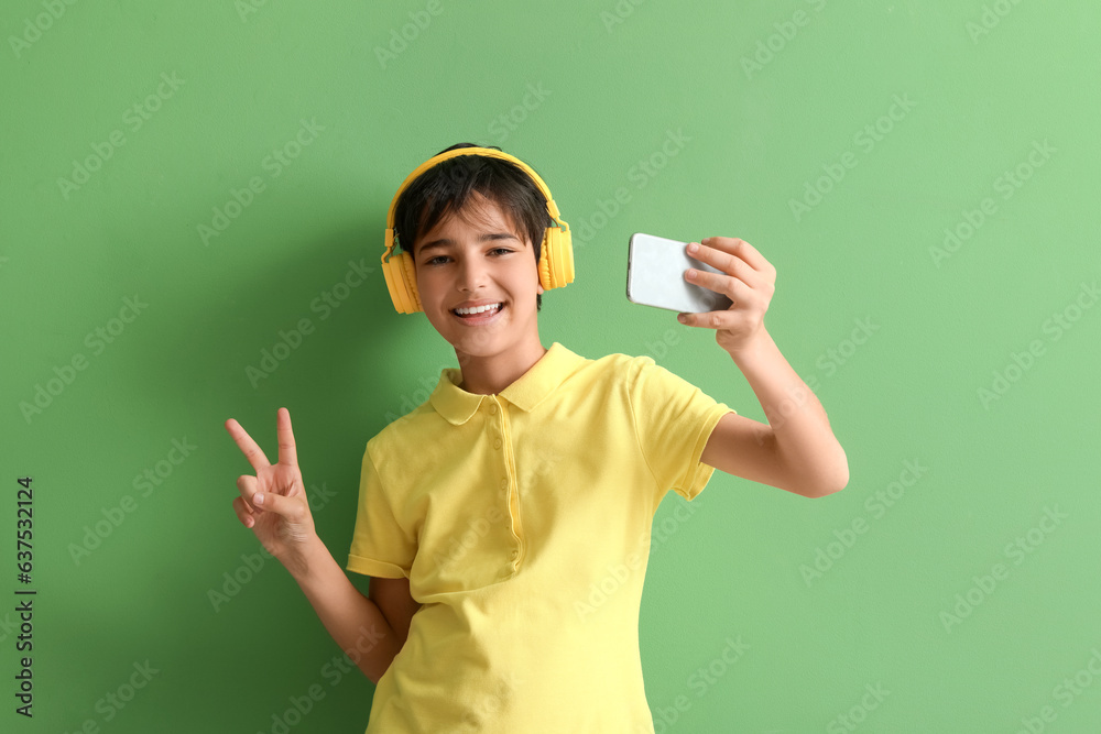 Little boy in headphones with mobile phone taking selfie on green background