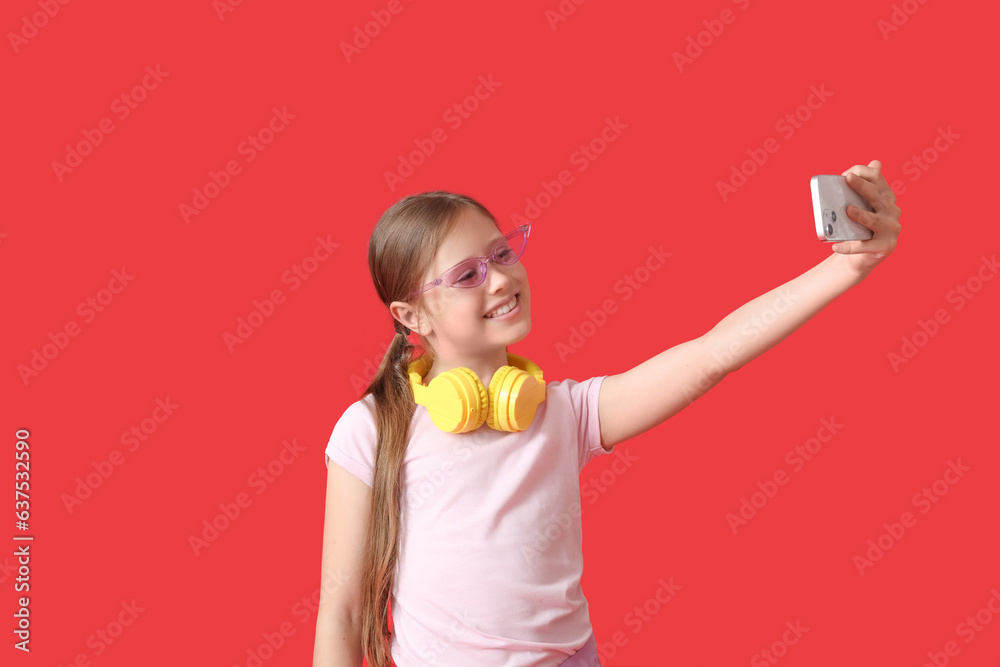 Little girl in headphones with mobile phone taking selfie on red background