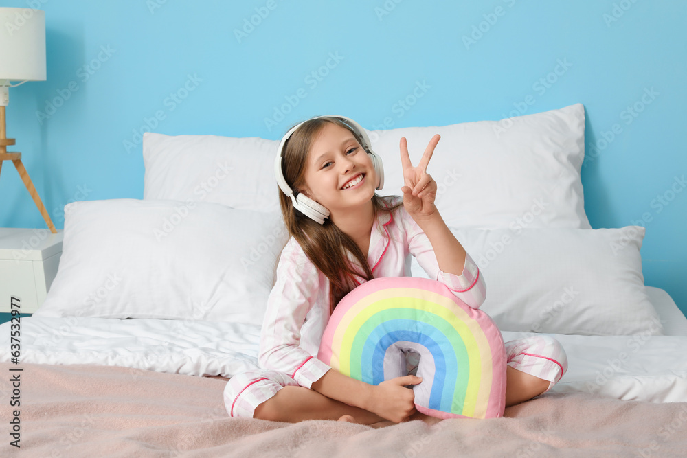 Little girl with headphones and rainbow pillow showing victory gesture in bedroom