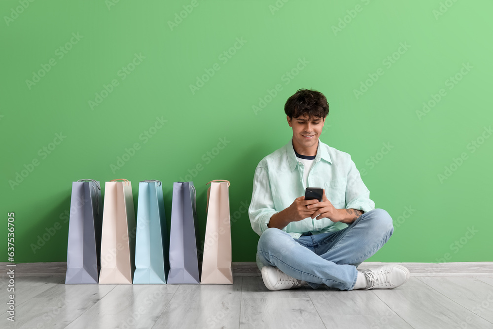 Young man with mobile phone and bags shopping online near green wall