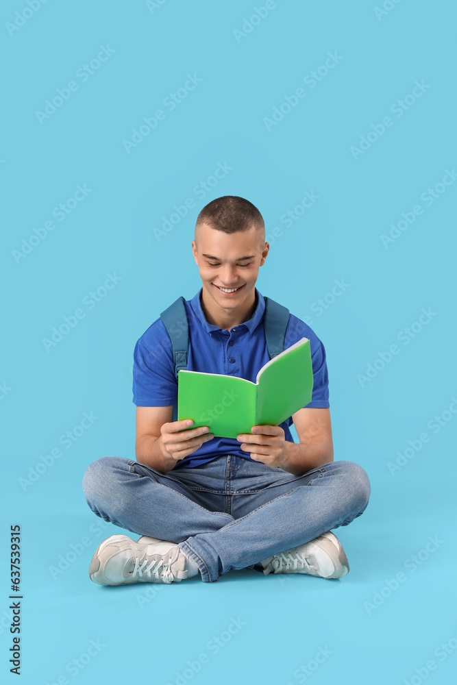 Happy male student with backpack sitting on blue background and reading book