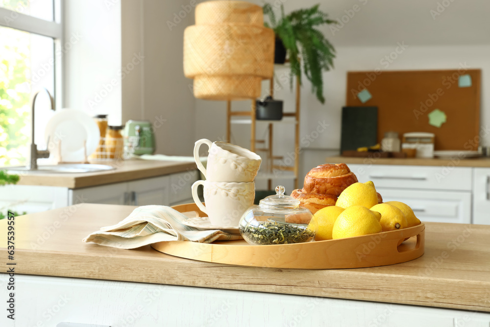 Tray with cinnamon rolls, cups, lemons and tea leaves on wooden countertop in modern kitchen