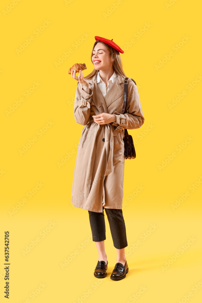 Beautiful stylish young woman in beret with tasty croissant on yellow background