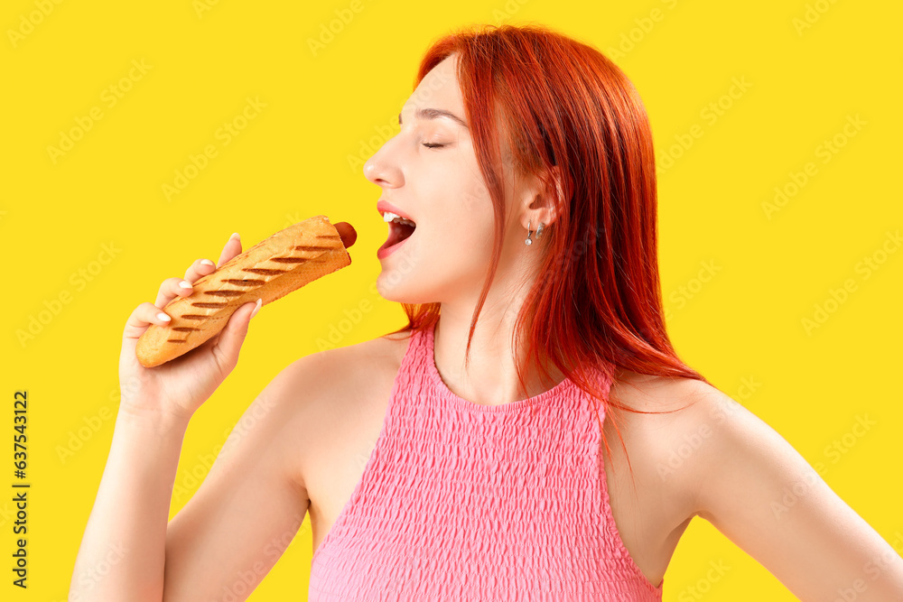 Beautiful young woman eating tasty french hot dog on yellow background