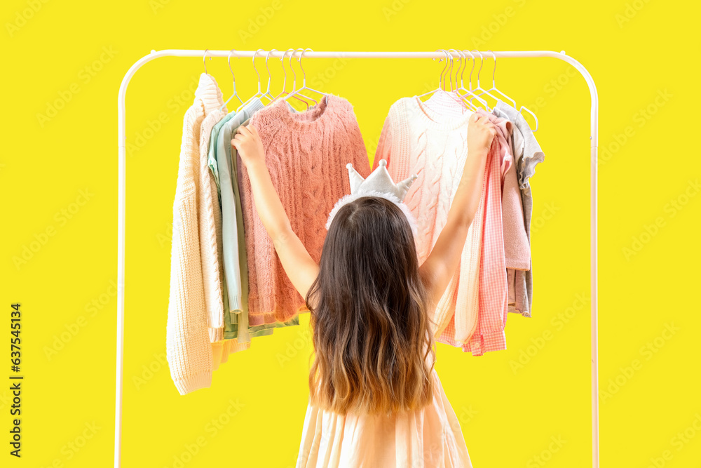 Cute little girl near rack with stylish clothes on yellow background, back view