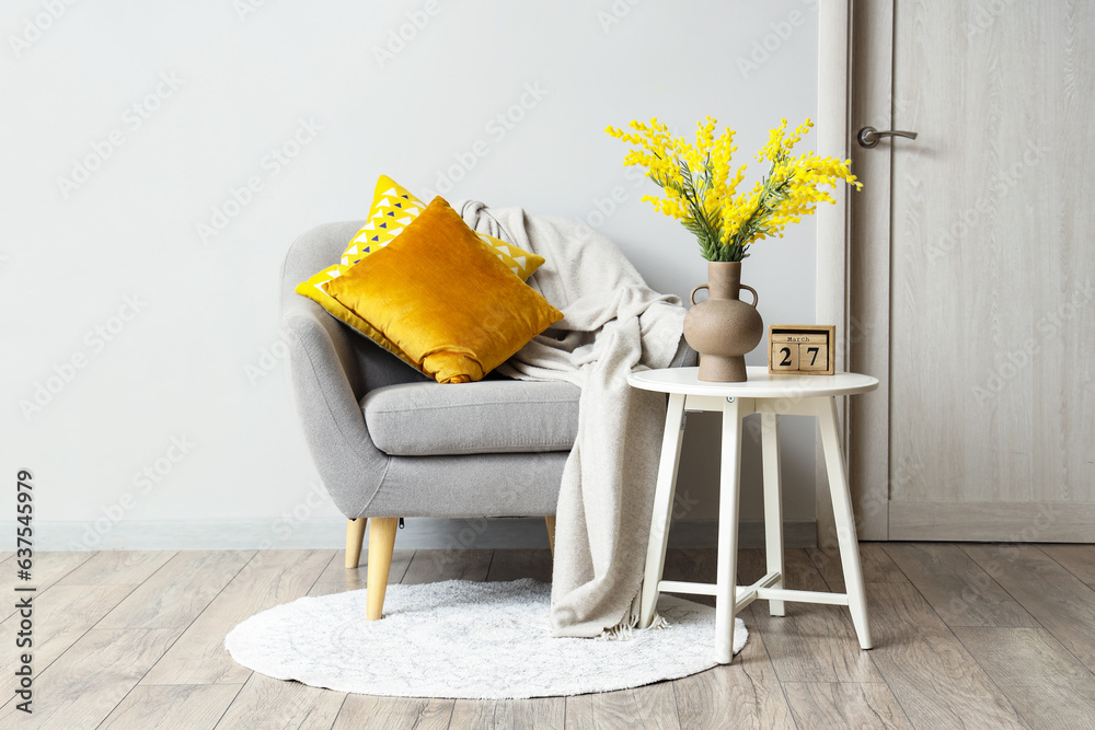 Vase with beautiful mimosa flowers on end table and comfortable armchair near white wall