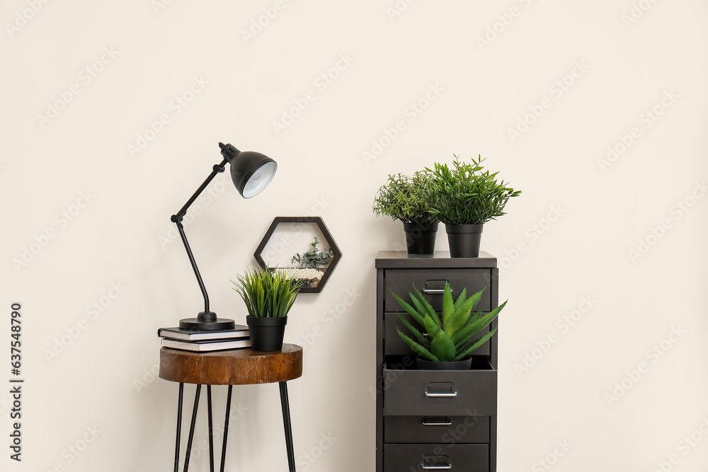 Desk lamp on small table and file cabinet with houseplants near white wall