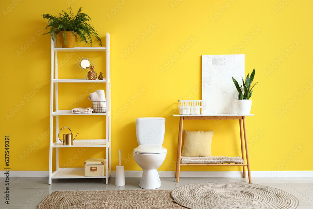 Interior of modern restroom with ceramic toilet bowl, shelving unit and console table near yellow wa