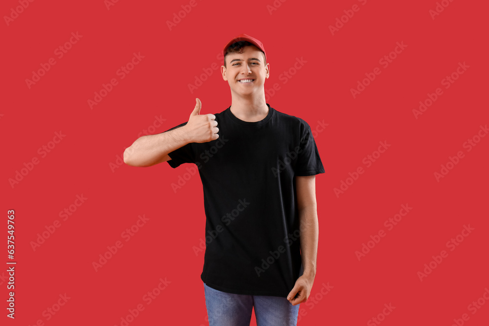 Young man in black t-shirt and cap showing thumb-up on red background