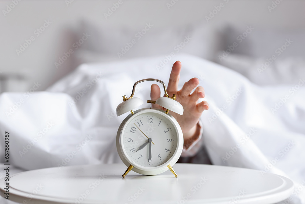 Female hand under blanket reaching for alarm clock in bedroom