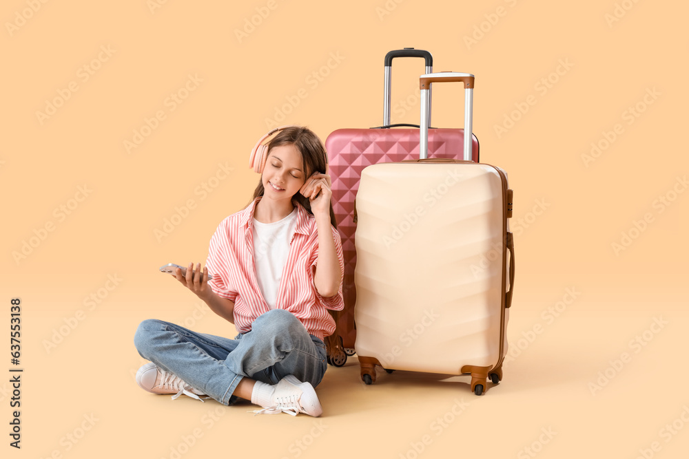 Little girl in headphones with mobile phone and suitcase sitting on orange background