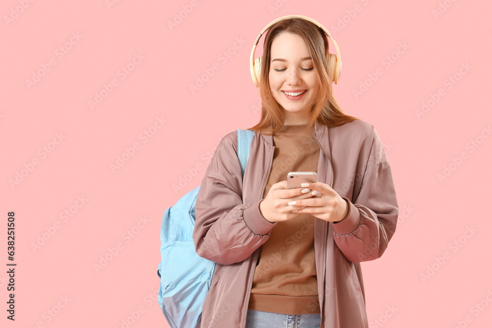 Female student in headphones using mobile phone on pink background