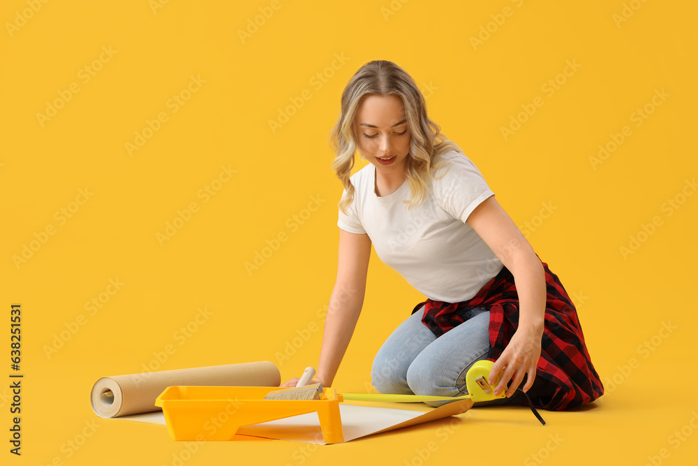 Young woman measuring wallpaper on yellow background