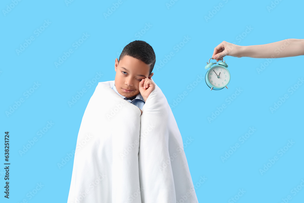 Sleepy little African-American boy and female hand with alarm clock on blue background