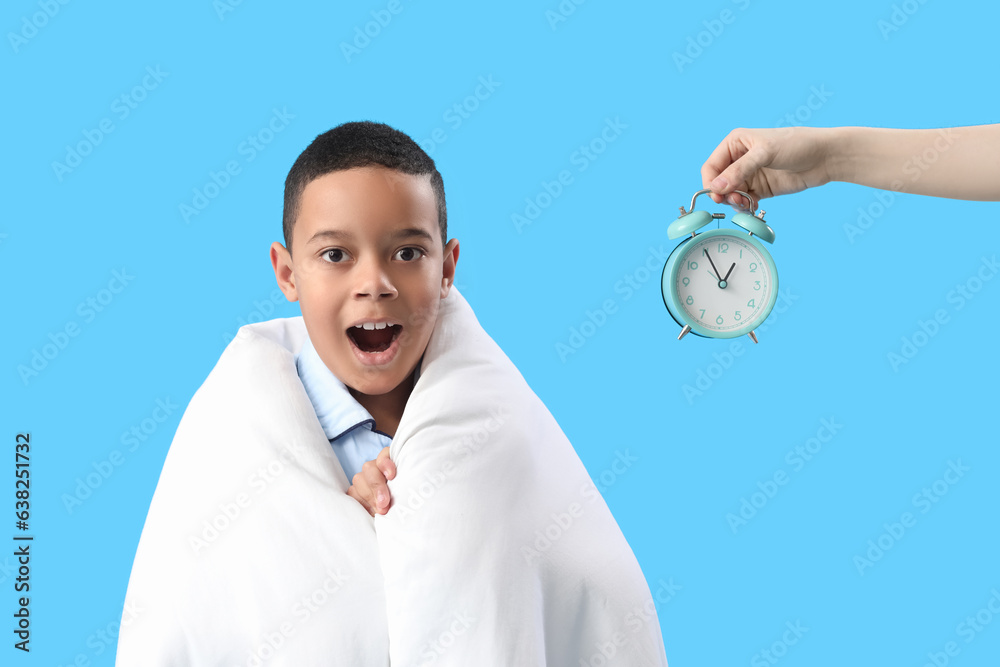 Surprised little African-American boy and female hand with alarm clock on blue background