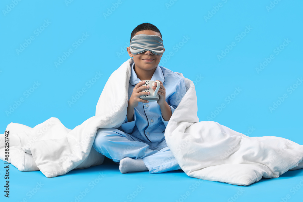 Little African-American boy in sleeping mask with cup of cocoa and soft blanket on blue background