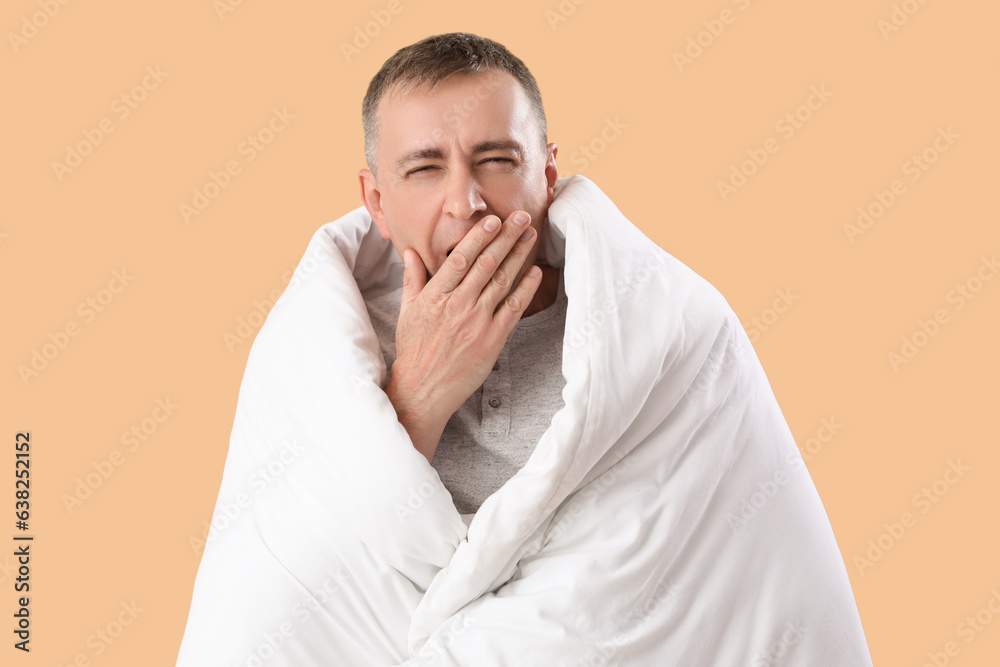 Mature man with soft blanket yawning on beige background