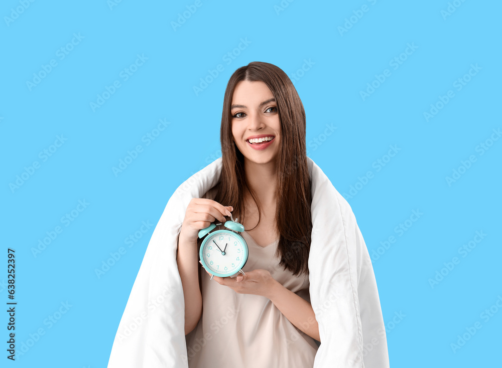 Young woman with alarm clock and soft blanket on blue background