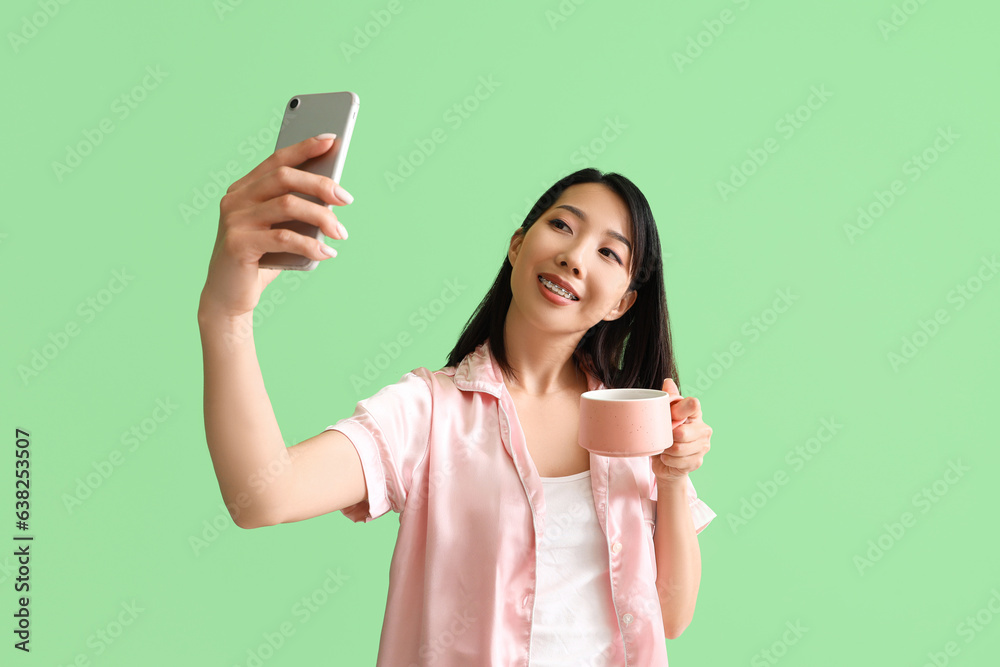 Beautiful Asian woman in pajamas with cup of coffee taking selfie on green background