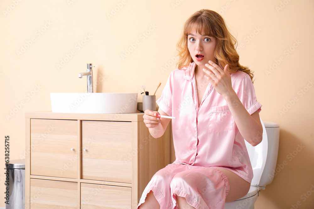 Shocked young woman with pregnancy test sitting on toilet bowl in restroom