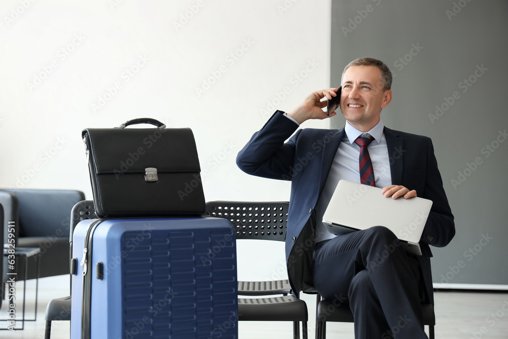 Mature businessman with laptop talking by mobile phone in hall of airport