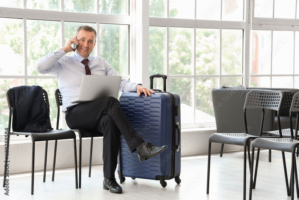 Mature businessman with laptop talking by mobile phone in hall of airport