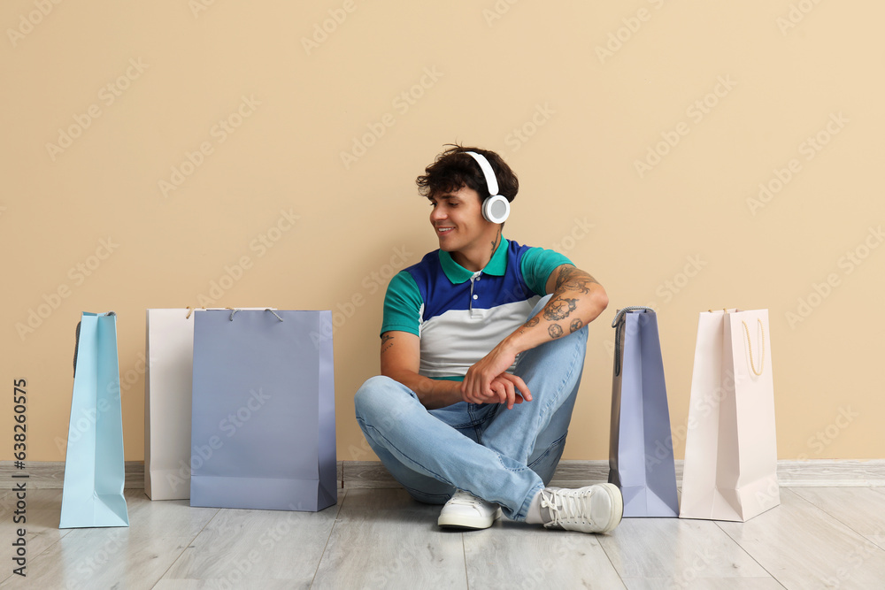 Young man in headphones with shopping bags sitting near beige wall