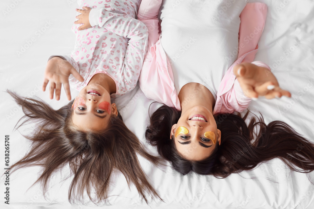 Happy mother and her little daughter with under-eye patches lying on bed, top view