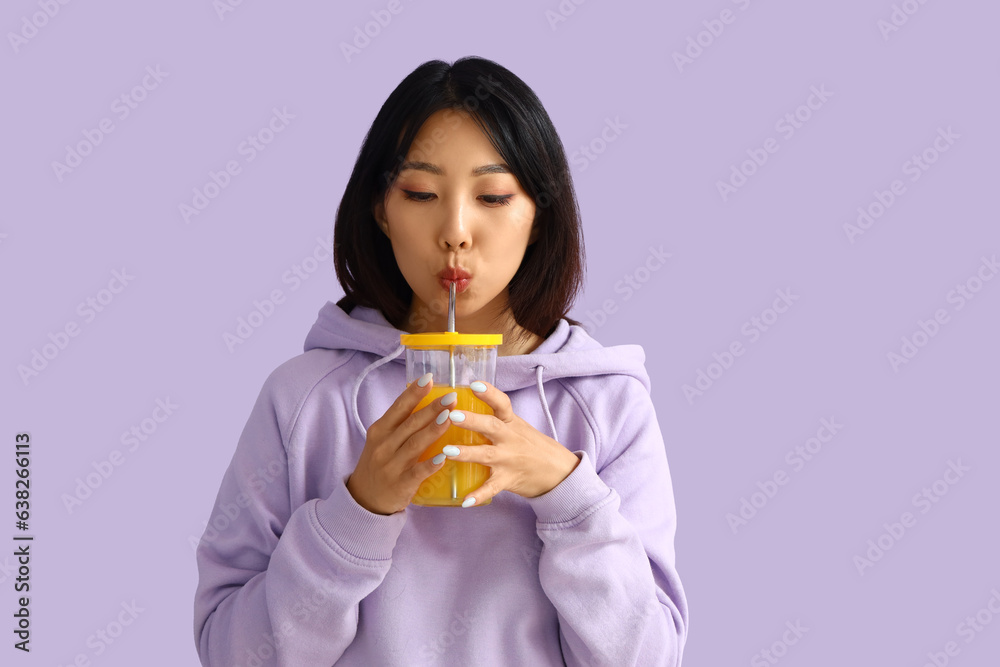 Beautiful Asian woman drinking juice on lilac background