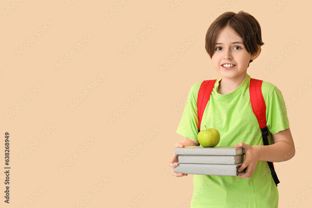 Happy little boy with backpack, books and apple on beige background