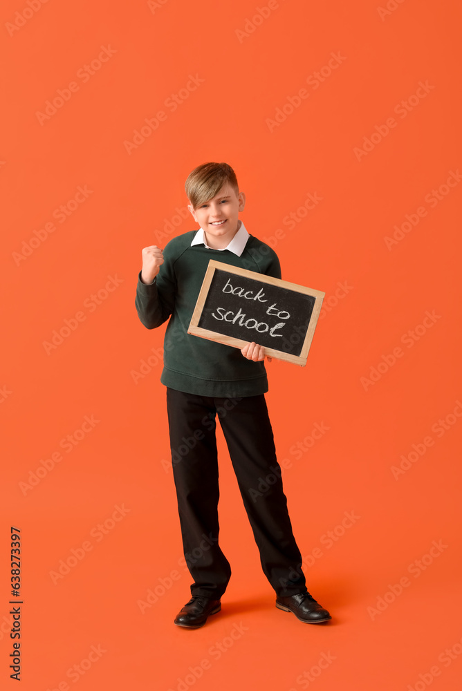 Little schoolboy in stylish uniform holding blackboard with text BACK TO SCHOOL on orange background