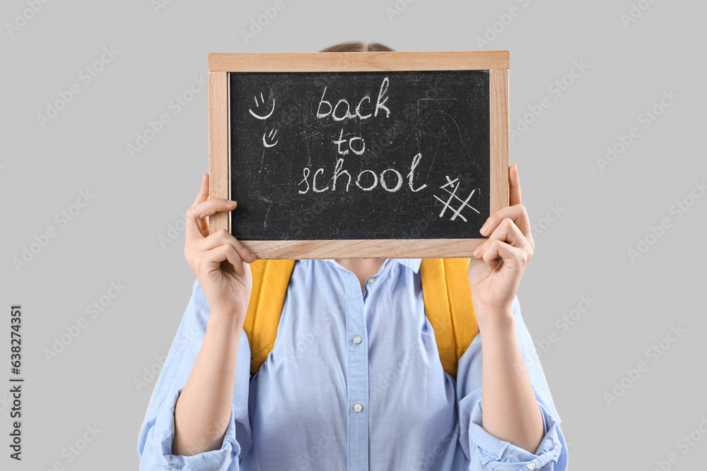 Schoolgirl in backpack holding blackboard with text BACK TO SCHOOL on grey background