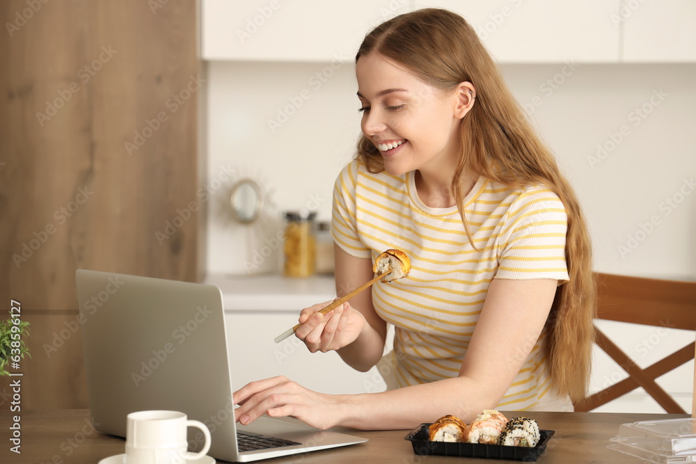 Happy young woman with laptop eating sushi in kitchen