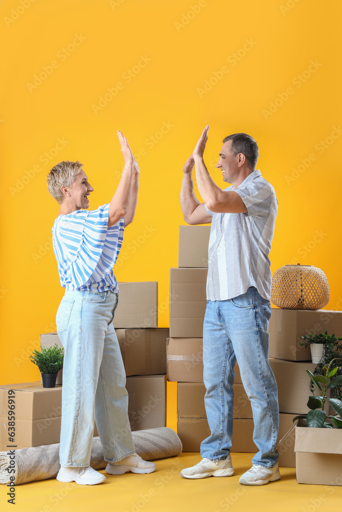 Mature couple with moving boxes giving each other high-five on yellow background