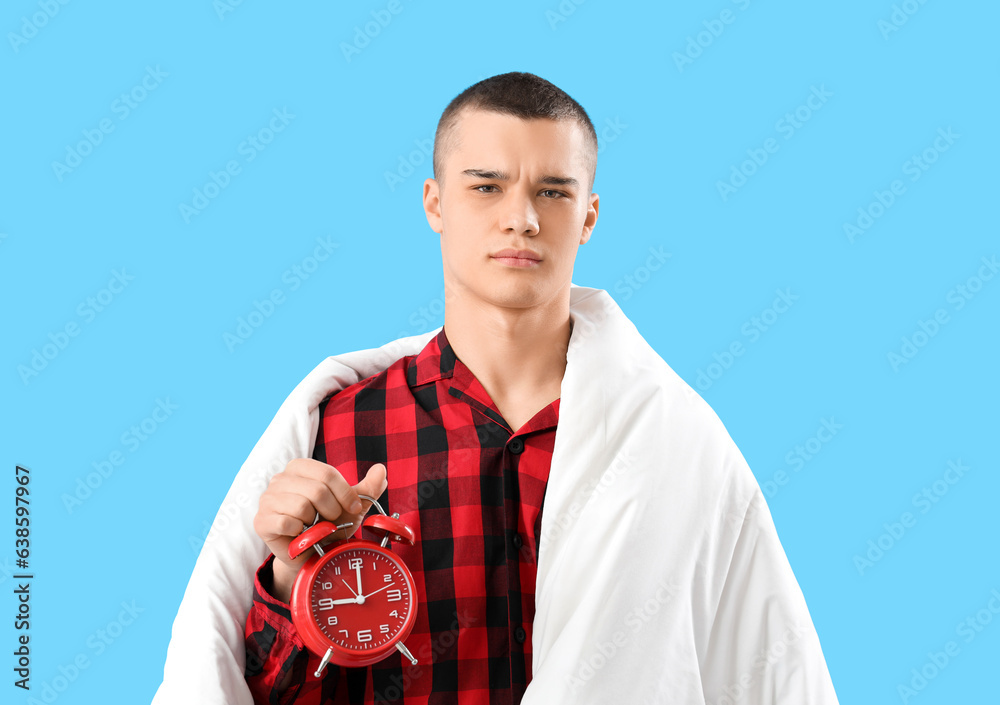 Displeased young man in pajamas, with blanket and alarm clock on blue background