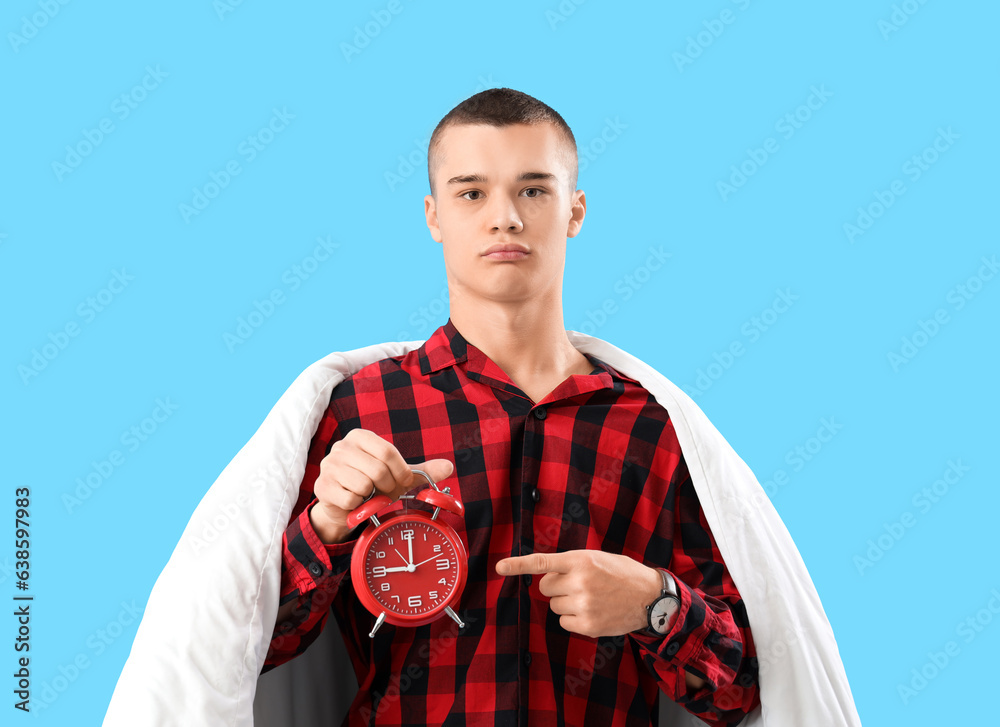 Young man in pajamas, with blanket and alarm clock on blue background