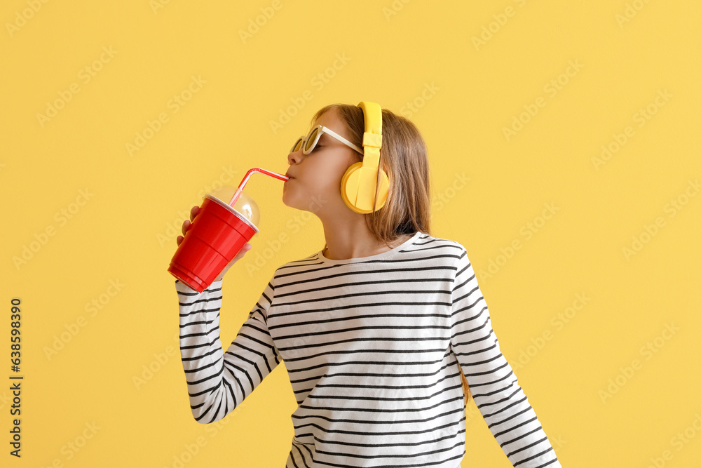 Little girl in headphones drinking soda on yellow background