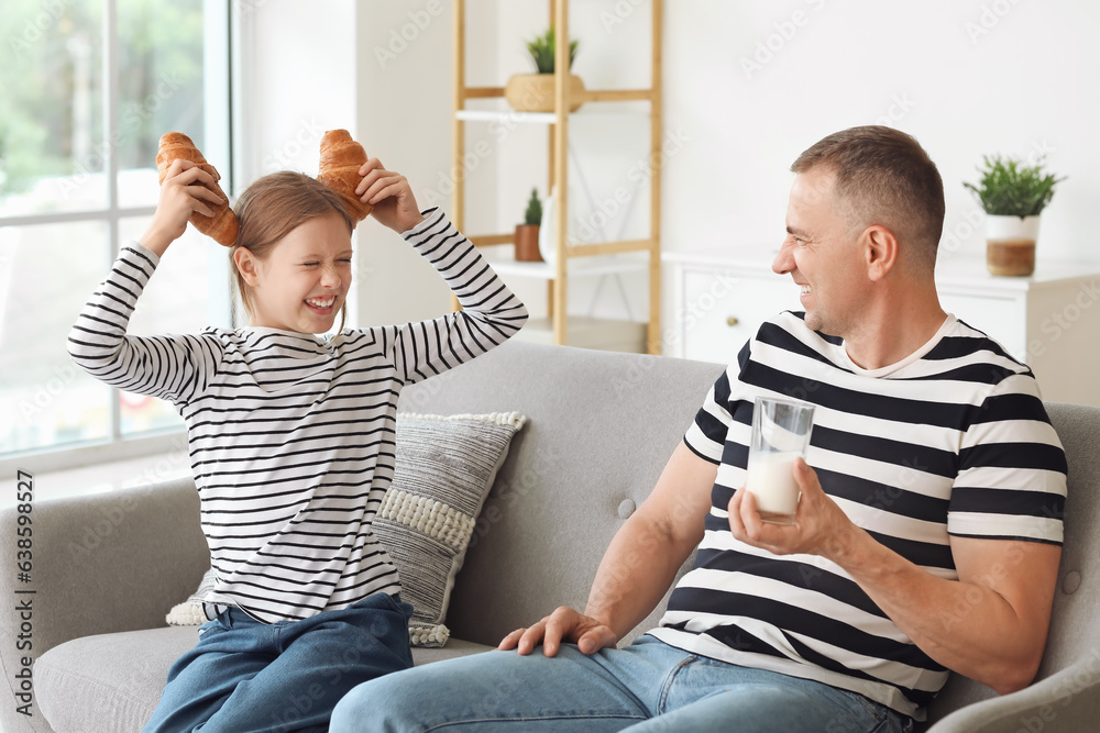 Little girl holding croissants with her father sitting on sofa and drinking milk in living room