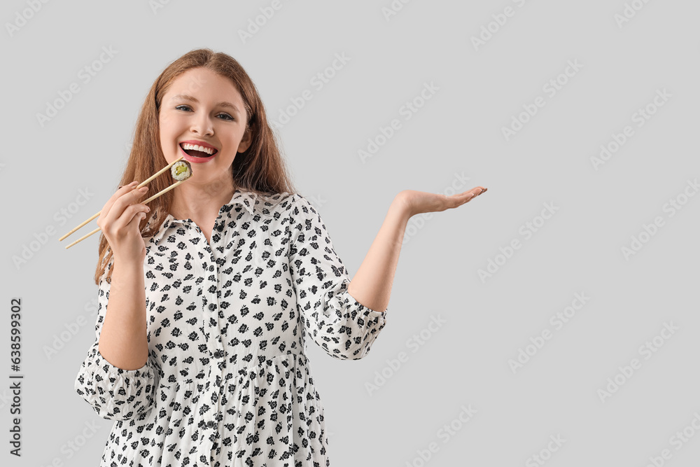 Young woman with tasty sushi roll showing something on grey background