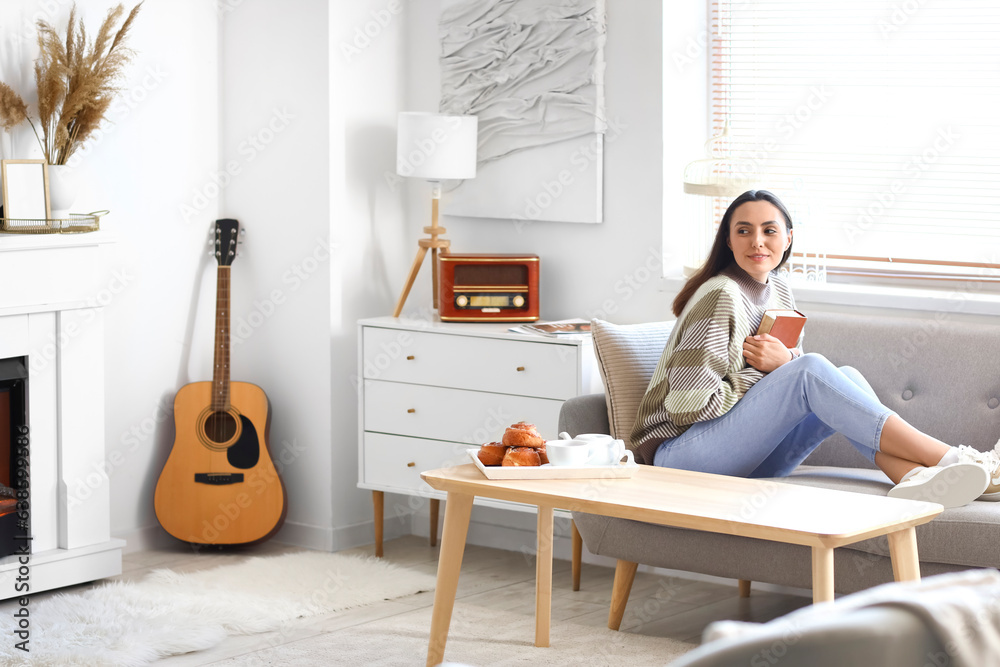 Young woman with book sitting on sofa near fireplace at home