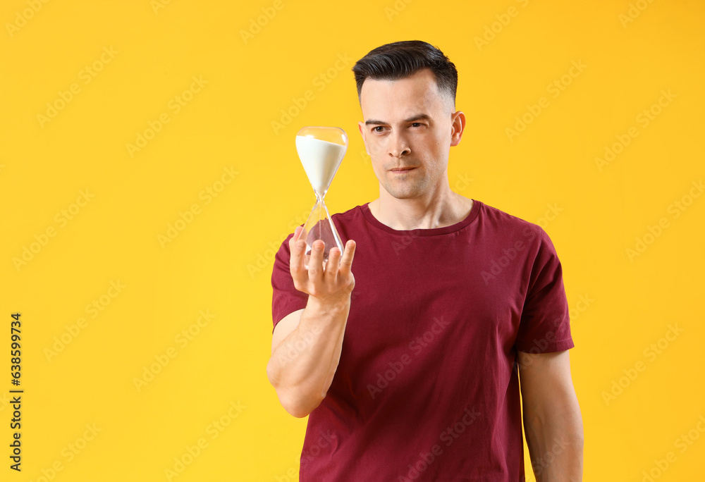 Stressed young man with hourglass on yellow background. Deadline concept