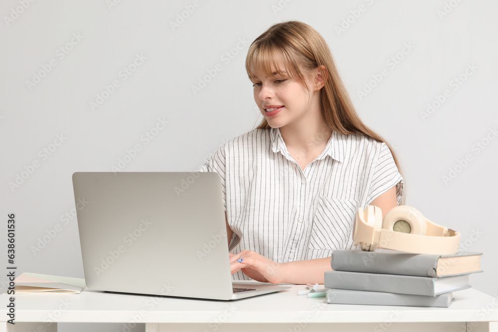 Female student with laptop doing homework at table on light background