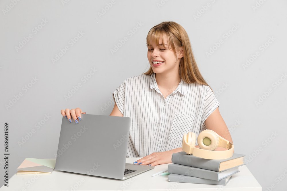 Female student with laptop doing homework at table on light background